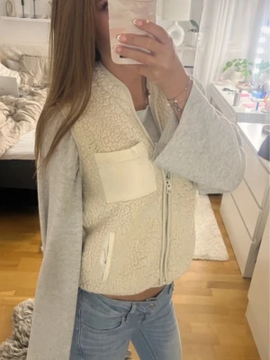 Beige teddy dunväst med fickor - Mysig beige teddy dunväst med dragkedja framtill och två fickor, en bröstficka och en sidoficka. Västen har en fluffig, fårullsliknande yta och är perfekt för lager-på-lager-stil. Passar till jeans och hoodie för en avslappnad look.