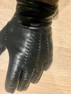 Svarta skinnhandskar med sömmar - Stilrena svarta handskar i skinn med dekorativa sömmar på ovansidan. Klassisk design med mjukt foder och diskret etikett vid handleden. Perfekta för kyliga dagar när du vill hålla händerna varma och snygga.