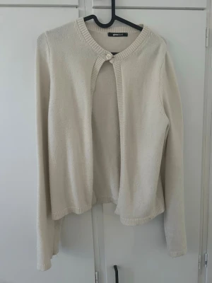Krämvit kofta från Gina Tricot - Stilren krämvit kofta från Gina Tricot med rund halsringning och en knapp framtill. Koftan har långa ärmar och ribbade kanter. Perfekt att slänga över en topp för en enkel och snygg look.            Vid knappen har koftan en liten skada men inget som syns särskild eller påverkar något.