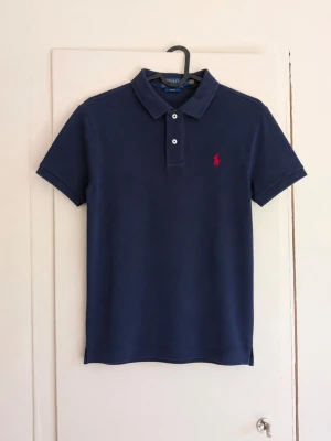Polo Ralph Lauren Slim Fit piké – L (14–16) 🐎 - Klassisk mörkblå pikétröja från Polo Ralph Lauren med röd broderad logga på bröstet. Slim Fit-modell med stilren och tidlös design.  🔹 Storlek: L (14–16 år) 🔹 Modell: Slim Fit 🔹 Färg: Mörkblå / navy 🔹 Röd broderad Polo-logo 🔹 Fint begagnat skick men den är har några missfärgningar! 🔹 QR-/produktetikett finns kvar  Passar perfekt både till vardags och lite finare tillfällen. Säljes då den inte längre kommer till användning.  Hör gärna av dig vid frågor eller funderingar!😁