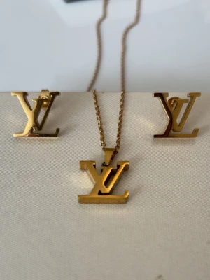 Guldfärgat smyckeset med LV-logga - Snyggt smyckeset med halsband och örhängen i guldfärg, båda med den ikoniska LV-loggan. Setet har en stilren och modern design som passar perfekt till att lyfta din outfit. Materialet är metall med blank finish.