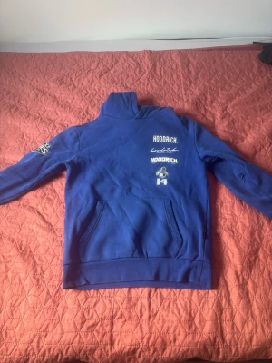 Blå hoodie från Hoodrich - Snygg blå hoodie från Hoodrich med vita tryck på bröstet och ärmen. Hoodien har en stor ficka framtill och en klassisk huva med tryck på insidan. Perfekt för dig som gillar streetwear och vill ha en bekväm och stilren look.