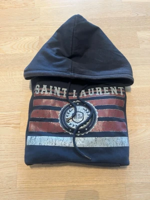 Svart Saint Laurent hoodie med tryck - Säljer en svart hoodie från Saint Laurent med stort tryck framtill i rött, vitt och blått. Hoodien har huva med dragsko och lång ärm. Trycket har texten 'SAINT LAURENT' och ett SL-emblem. Perfekt för dig som gillar streetwear och snygga detaljer.