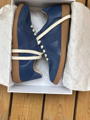 Blå Margiela’s sneakers - Säljer snygga maison margiela blå sneakers med ovandel i både mocka och slätt läder. Skorna har vita snören och en klassisk beige gummisula som ger retrovibbar. Perfekta för dig som vill ha en stilren men ändå unik look till vardagsoutfiten. 