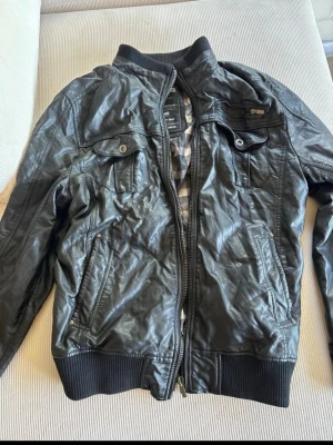 Svart skinnjacka med dragkedja - Snygg svart skinnjacka med dragkedja framtill, ribbade muddar vid ärmslut och midja samt flera fickor med både dragkedja och knappar. Jackan har en klassisk bomberinspirerad look och rutigt innerfoder.