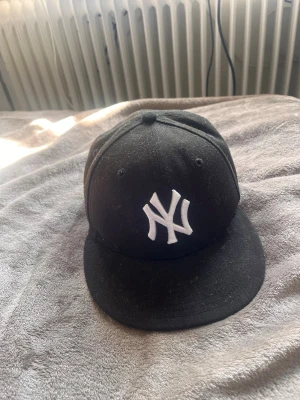 Svart New York Yankees keps - Säljer en svart keps från New Era med klassisk New York Yankees-logga broderad i vitt framtill. Kepsen har rak skärm och broderade ventilationshål. Perfekt för dig som gillar streetwear och sportig stil.