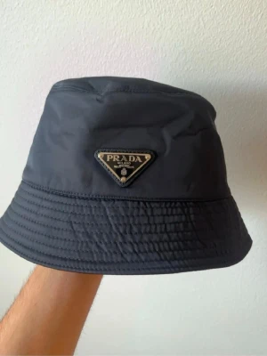 Svart bucket hat från Prada - Snygg svart bucket hat från Prada i nylonmaterial med det klassiska trekantiga metallogot på framsidan. Hatten har en stilren och modern look som passar perfekt till streetwear och trendiga outfits.