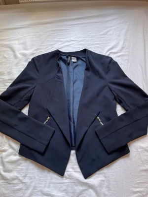 Mörkblå kavaj från H&M Divided - Stilren mörkblå kavaj från H&M Divided med öppen front och två snedställda dragkedjefickor. Kavajen har en mjuk, slät yta och helfodrad insida i matchande blått. Perfekt för en trendig och enkel look.