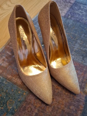 Beige glittriga pumps från Catwalk - Säljer ett par snygga pumps från Catwalk i beige med glittrig yta och diskret mönster. Skorna har spetsig tå och hög klack, samt guldfärgad innersula som ger en lyxig känsla. Perfekta för dig som vill sticka ut lite extra.