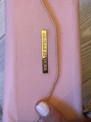 Ljusrosa plånbok från Ideal of Sweden💕 - Snygg ljusrosa plånbok/mobilskal från Dela of Sweden med guldfärgad logga på framsidan. Plånboken/mobilskalet har en stilren design med mjukt tygmaterial och smart knappstängning. Perfekt för dig som gillar en feminin och trendig accessoar.