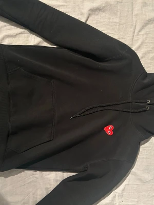 Svart hoodie med rött hjärta  - Säljer en svart hoodie med huva och känguruficka framtill. På bröstet sitter en liten röd hjärtdetalj med ögon som ger en unik touch. Märket är comme des Garcons och är i stolek M. Pris kan såklart diskuteras🤍
