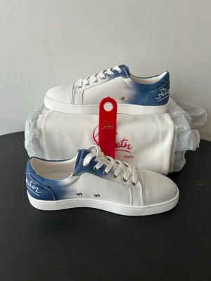 Vita och blå sneakers från Louboutin - Snygga sneakers från Christian Louboutin i vitt tyg med blå detaljer och logga på hälen. Skorna har klassisk låg profil, vita snören och den ikoniska röda sulan. Perfekta för dig som vill ha en stilren men ändå unik look. Allt OG medföljer förutom OG box