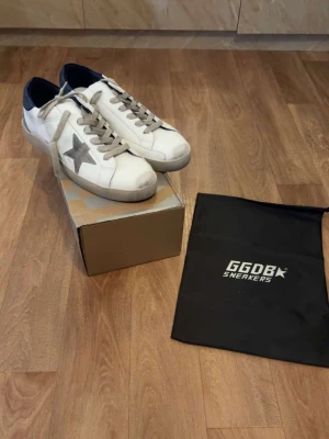 Vita GGDB sneakers med grå stjärna - Säljer ett par vita Golden Goose sneakers med grå stjärna på sidan och grå snören. Skorna har en klassisk låg silhuett och detaljer i mocka samt svart hälparti. Kommer med originalpåse. Perfekta för dig som gillar streetstyle och unika detaljer.