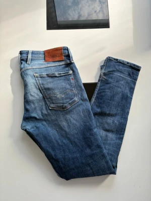 Blå slitna jeans med hål - Säljer ett par blå jeans med slitningar och hål på framsidan. Jeansen har klassisk femficksdesign, normal midja och raka ben. Materialet är denim i bomull och de har en brun patch bak i midjan. Perfekta för en avslappnad och trendig look.