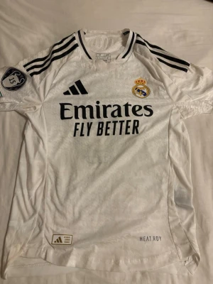 Vit Real Madrid t-shirt Adidas - Säljer en vit Real Madrid t-shirt från Adidas med svarta detaljer och klubbmärke på bröstet. Tröjan har korta ärmar, tre svarta ränder på axlarna och tryck med 'Emirates Fly Better' på framsidan. Perfekt för fotbollsfans!