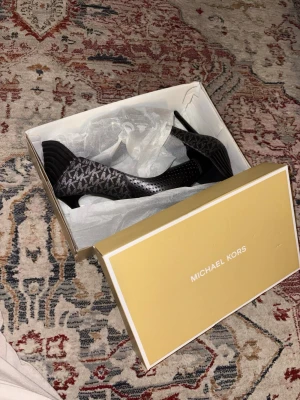 Svarta pumps från Michael Kors - Snygga svarta pumps från Michael Kors med spetsig tå och diskreta silverprickar framtill. Sidan har det klassiska MK-mönstret i grått och svart. Skorna är i textil och läder, perfekta för dig som vill ha en trendig och elegant look.