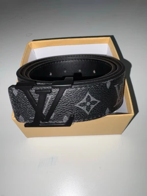 Svart bälte i skinn - Stilrent svart bälte från med klassiskt monogrammönster och stor logga i metall som spänne. Tillverkat i skinn med detaljer i svart och grått. Perfekt accessoar för att lyfta din outfit.