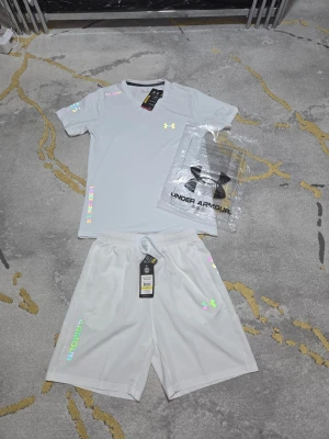 Vitt träningsset från Under Armour - Snyggt vitt träningsset från Under Armour med t-shirt och shorts. Setet har färgglada loggor och detaljer, samt UA-tryck på ärmen och benet. Tillverkat i ett lätt och snabbtorkande material som passar perfekt för träning och sport.