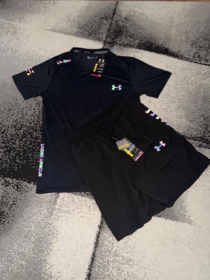 Svart träningsset från Under Armour - Säljer ett svart träningsset från Under Armour med t-shirt och shorts. Setet har färgglada loggor och textdetaljer på både tröja och byxa. Perfekt för gymmet eller löpning. Materialet är lätt och andas bra.