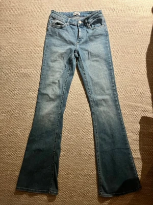 Blå bootcut jeans - Säljer ett par blå bootcut jeans med klassisk femficksdesign. Jeansen har en lätt tvättad look och vida ben från knät och ner. Perfekta för en avslappnad och trendig stil. Materialet är denim i bomull.