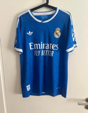 Blå Real Madrid matchtröja Adidas - Blå Real Madrid matchtröja från Adidas med vita detaljer och klubbmärke på bröstet. Tröjan har rund hals, tryckt sponsorlogga och klassiska Adidas-ränder på axlarna. Perfekt för fotbollsträning eller att bära till vardags.
