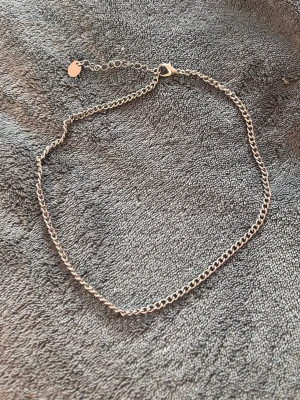 Silver kedja  - Säljer ett stilrent och tunt halsband i silverfärg med enkel kedjedesign. Halsbandet har ett justerbart lås och en liten rund bricka vid förlängningskedjan. Perfekt för att kombinera med andra smycken eller bära solo.