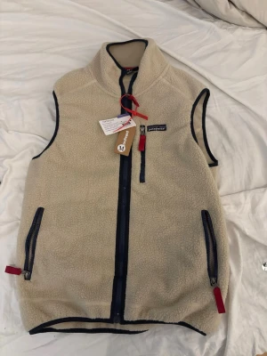 Beige fleeceväst från Patagonia - Snygg beige fleeceväst från Patagonia med svarta detaljer och röda dragkedjor. Västen har hög krage, tre fickor med dragkedja och är tillverkad i mjukt teddyfoder. Perfekt för lager-på-lager och outdoor-stil.