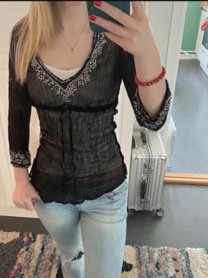 Y2K Sheer Black Beaded Top - This is a sheer, black, crinkled mesh top with white beaded floral embellishments around the neckline and sleeve cuffs. It has a deep V-neck and 3/4 bell sleeves. The top is fitted and semi-transparent, perfect for layering over a tank or bralette. The style is reminiscent of early 2000s (Y2K) fashion, often seen in brands like Free People, Express, or vintage boutiques. No visible brand tag in the provided images.
