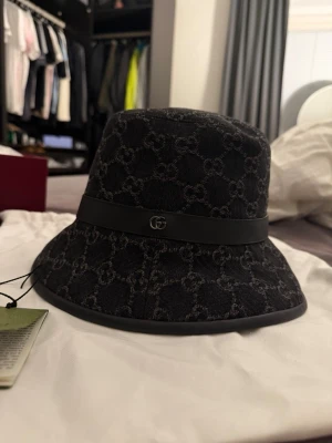 Svart bucket hat från Gucci - Snygg svart bucket hat från Gucci med det klassiska GG-monogrammet i grått över hela hatten. Hatten har ett svart band runtom med en liten metallogo framtill. Tillverkad i tyg med exklusiv känsla och trendig form.