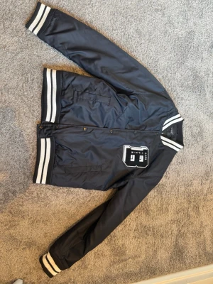 Svart bomberjacka med vita detaljer - Snygg svart bomberjacka från The Black Label Collection med vita ränder på krage, ärmslut och nederkant. Jackan har tryckknappar, två fickor och en stor vit patch på bröstet. Materialet är glansigt och ger en sportig collegekänsla.