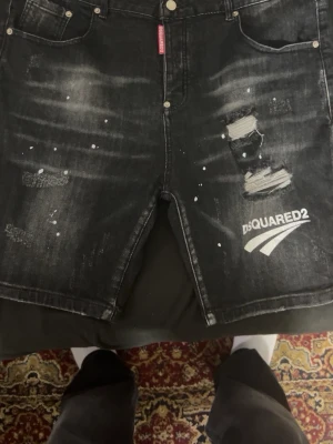 Svarta jeansshorts från Dsquared2 - Säljer ett par svarta jeansshorts från Dsquared2 med slitningar, tryckt logga och coola detaljer som färgstänk och trasiga partier. Klassisk femficksmodell med knappgylf och en relaxed fit. Perfekta för dig som gillar streetwear och vill sticka ut.