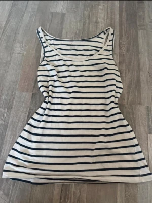 Randig vit och blå linnetopp - Säljer en vit linnetopp med marinblå horisontella ränder. Toppen är ärmlös och har en rundad halsringning. Perfekt till jeans eller shorts för en avslappnad look.