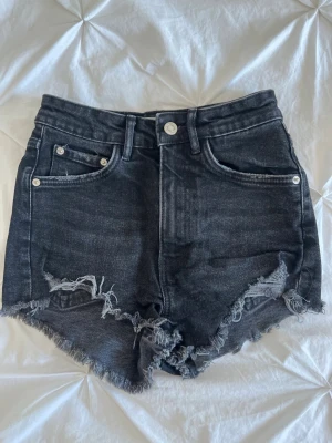 Svarta högmidjade jeansshorts - Säljer ett par svarta högmidjade jeansshorts med råa, fransiga kanter och klassisk femficksdesign. Shortsen har en mörk tvätt och silverfärgade nitar och knapp. Perfekta för sommaren och ger en cool, avslappnad vibe.