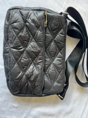 HVISK: Svart quiltad axelväska - Snygg svart axelväska med quiltat mönster och justerbar axelrem. Väskan har dragkedja framtill och ett prickigt innerfoder med innerfack och dragkedja. Perfekt för dig som vill ha en stilren och praktisk väska till vardagen. 22x15 cm