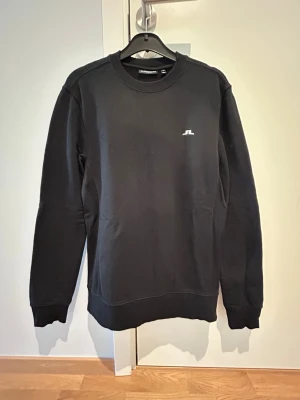 Svart sweatshirt från J.Lindeberg - Stilren svart sweatshirt från J.Lindeberg med rund halsringning och diskret logga på bröstet. Tröjan har långa ärmar, ribbade muddar och är gjord i mjukt bomullsmaterial. Perfekt för en clean och avslappnad look.