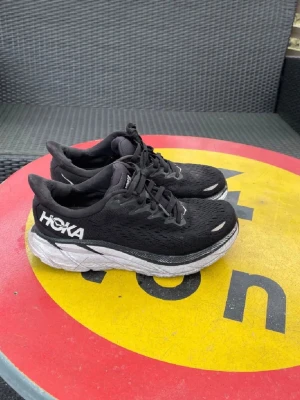 Svarta Hoka sneakers med tjock sula - Säljer ett par svarta Hoka sneakers med vit och svart tjock sula. Skorna har ventilerande mesh-överdel, klassisk snörning och Hoka-logga i vitt på sidan. Perfekta för dig som gillar sportig stil och komfort.