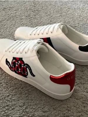 Gucci vita sneakers med orm - Snygga vita sneakers från Gucci med ikonisk orm-brodyr på sidan och klassiska gröna/röda detaljer. Baksidan har metallicröd och metallicgrön hälflik med Gucci-logga. Skorna är låga och har vita snören samt gummisula. Perfekt för dig som gillar statement-plagg.