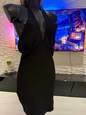 Svart halterneck klänning - Snygg svart halterneck klänning med djup urringning och figurnära passform. Klänningen är kort och ärmlös, perfekt för dig som vill ha en stilren och enkel look. Materialet ser ut att vara mjukt och stretchigt, vilket ger en bekväm känsla.