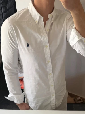 Vit skjorta från Ralph Lauren - Klassisk vit skjorta från Ralph Lauren med button-down krage och broderad marinblå logga på bröstet. Skjortan har långa ärmar, knäppning framtill och är tillverkad i bomull. Perfekt för en stilren och tidlös look.