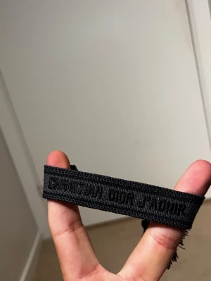 Svart armband Christian Dior - Snyggt svart armband från Christian Dior med broderad text i samma färg. Armbandet är gjort i tyg och har justerbara tofsar i ändarna för en cool och avslappnad look. Perfekt accessoar för att lyfta din outfit.