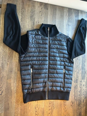 Svart cardigan från icon - Snygg svart cardigan med quiltat mönster och hel dragkedja framtill. Västen har två dragkedjeförsedda fickor och stickade svarta ärmar samt muddar. Materialet är en mix av dun och stickat tyg vilket ger en cool kontrast och modern look.