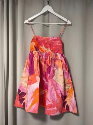 Sommarklänning - Säljer en kort klänning med tunna axelband och färgstarkt blommigt mönster i rosa, orange, lila och vitt. Klänningen har en luftig passform och är perfekt för varma dagar. Materialet känns lätt och bekvämt, och modellen ger en härlig sommarkänsla. Perfekt för kräftskiva 🦞🦞
