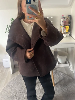 Mörkbrun ullkappa med bred krage - Snygg mörkbrun kappa i ullmix med oversized passform och bred krage. Kappan har knäppning framtill med stora knappar och raka ärmar. Perfekt för kyliga dagar och ger en stilren look till alla outfits.