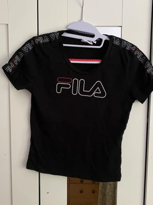 Svart FILA t-shirt med logga - Snygg svart t-shirt från FILA med stor logga framtill och logoband längs axlarna. T-shirten har rund halsringning och korta ärmar. Perfekt för en sportig och trendig look.