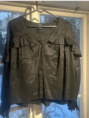 Svart blus med volanger - Snygg svart blus från Lindex med glansigt tyg och volangdetaljer över axlarna. Blusen har knappar framtill och v-ringning. Perfekt för dig som gillar en trendig och elegant look. Materialet ser ut att vara polyester eller liknande.