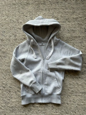 Ljusgrå zip hoodie russemerch - Säljer en stilren ljusgrå hoodie med dragkedja och stor huva. Tröjan har fickor framtill, ribbade muddar vid ärmar och nederkant samt dragsko i huvan. Perfekt för en chill och avslappnad look. Materialet är mjukt och skönt, troligtvis bomullsblandning.
