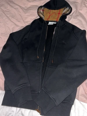 Svart hoodie med dragkedja och huva - Svart hoodie med dragkedja och huva som har ett klassiskt rutigt mönster på insidan. Tröjan har långa ärmar, fickor framtill och justerbara snören i huvan. Perfekt för en chill och avslappnad stil.
