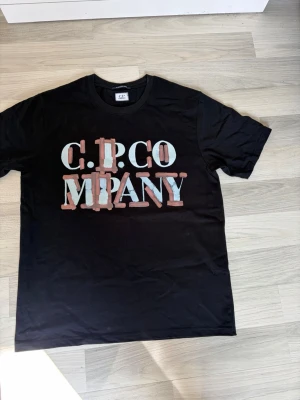 Svart C.P. Company t-shirt - Snygg svart t-shirt från C.P. Company med stor grafisk logga i vitt och ljusblått med rosa detaljer på bröstet. Klassisk rund hals och korta ärmar. Perfekt till jeans eller shorts för en avslappnad look.