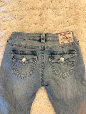 Joey lowrise True Religion - Säljer ett par ljusblå bootcut jeans från True Religion med ikoniska ficklock och vita kontrastsömmar. Jeansen har silverfärgade knappar och patch med logga bak i midjan. Klassisk femficksmodell i mjuk denim.