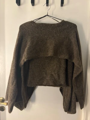 Brun stickad tröja med cape - Unik brun stickad tröja med cape. Tröjan har rund halsringning och är gjord i ett mjukt, ull-liknande material. Perfekt för dig som gillar en avslappnad och trendig stil.